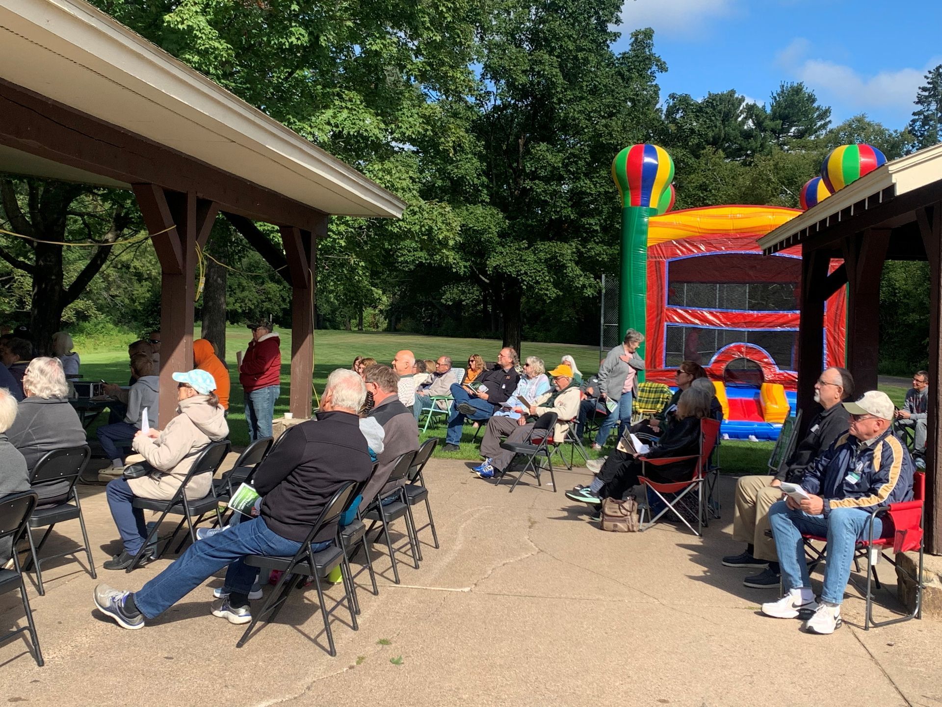 People seated outdoors at a park gathering near a colorful inflatable bounce house and pavilion.