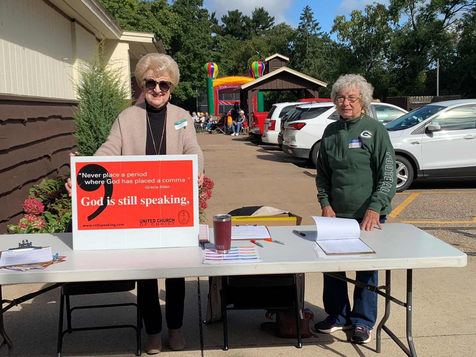 Two women stand behind a table with a red sign reading “God still speaks,” outdoors near parked cars.