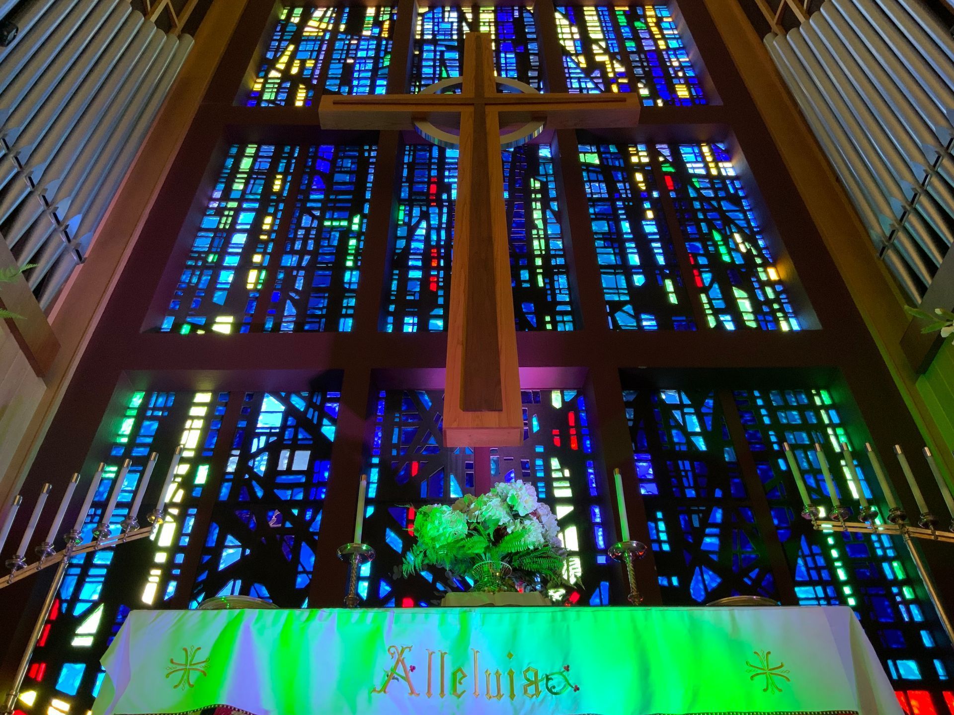 Stained-glass church windows above an altar with a green floral arrangement and cross.