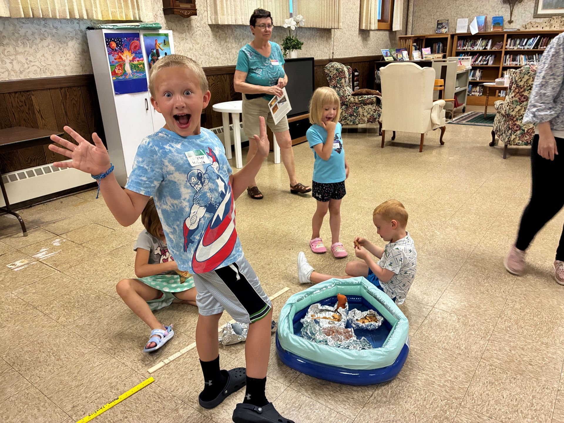Children playing in a room, with a boy waving beside a small blue inflatable pool.