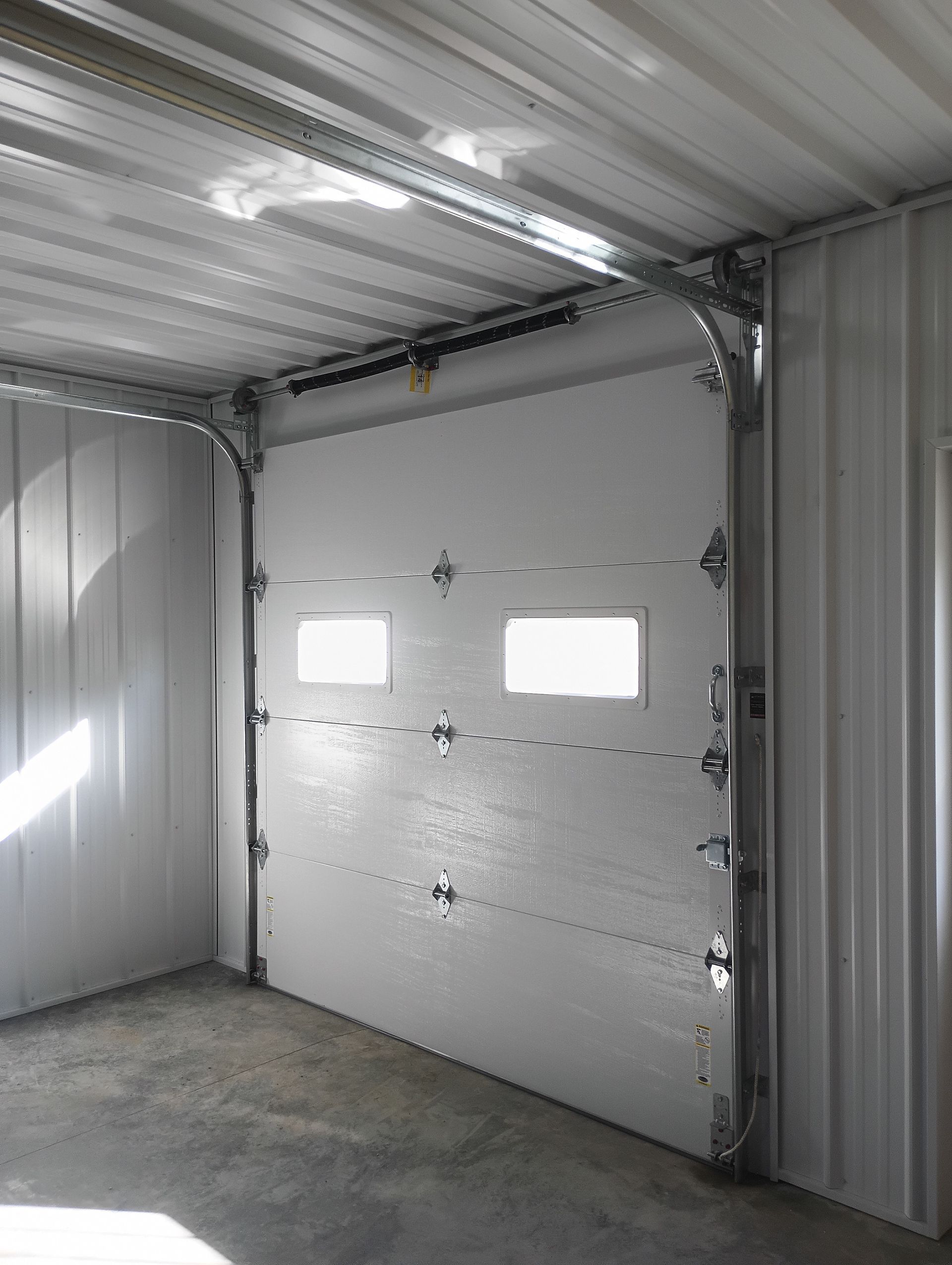 White industrial garage door, partially open, with two windows, inside a metal building.