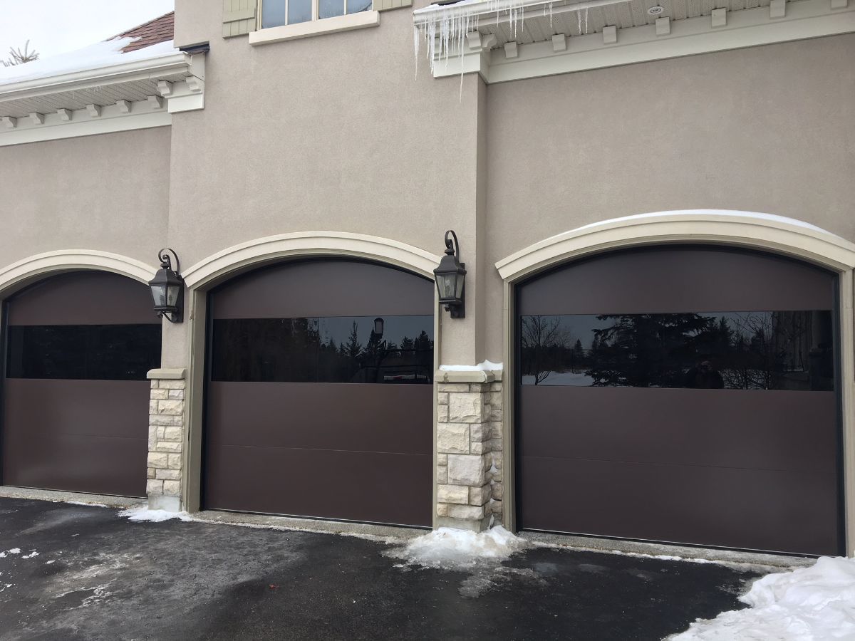 Three brown garage doors with a dark tinted window and stone accents, in a snowy setting.