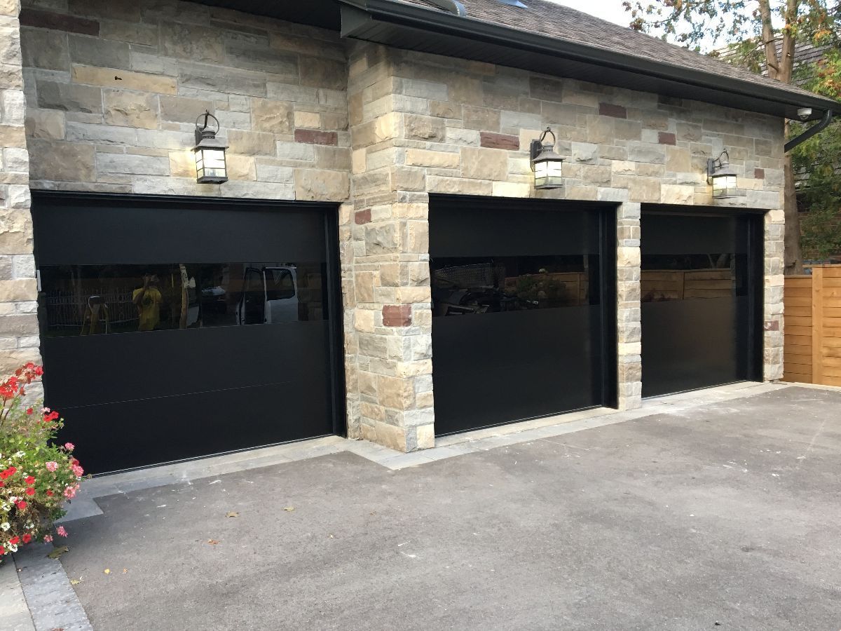 Three-car garage with black doors, stone facade, and outdoor lights. Asphalt driveway with plants on the left.