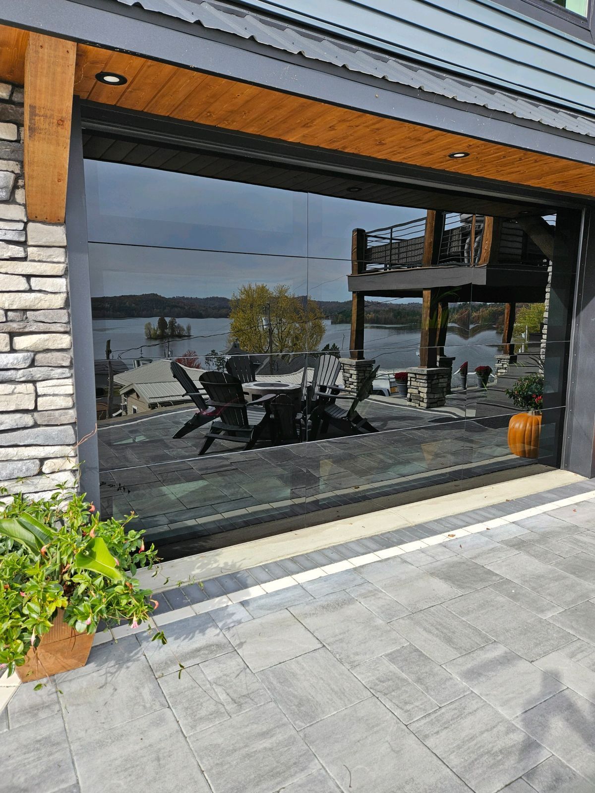 Large glass window reflects outdoor patio furniture, lake, and a deck. Stone exterior, wooden beam roof.