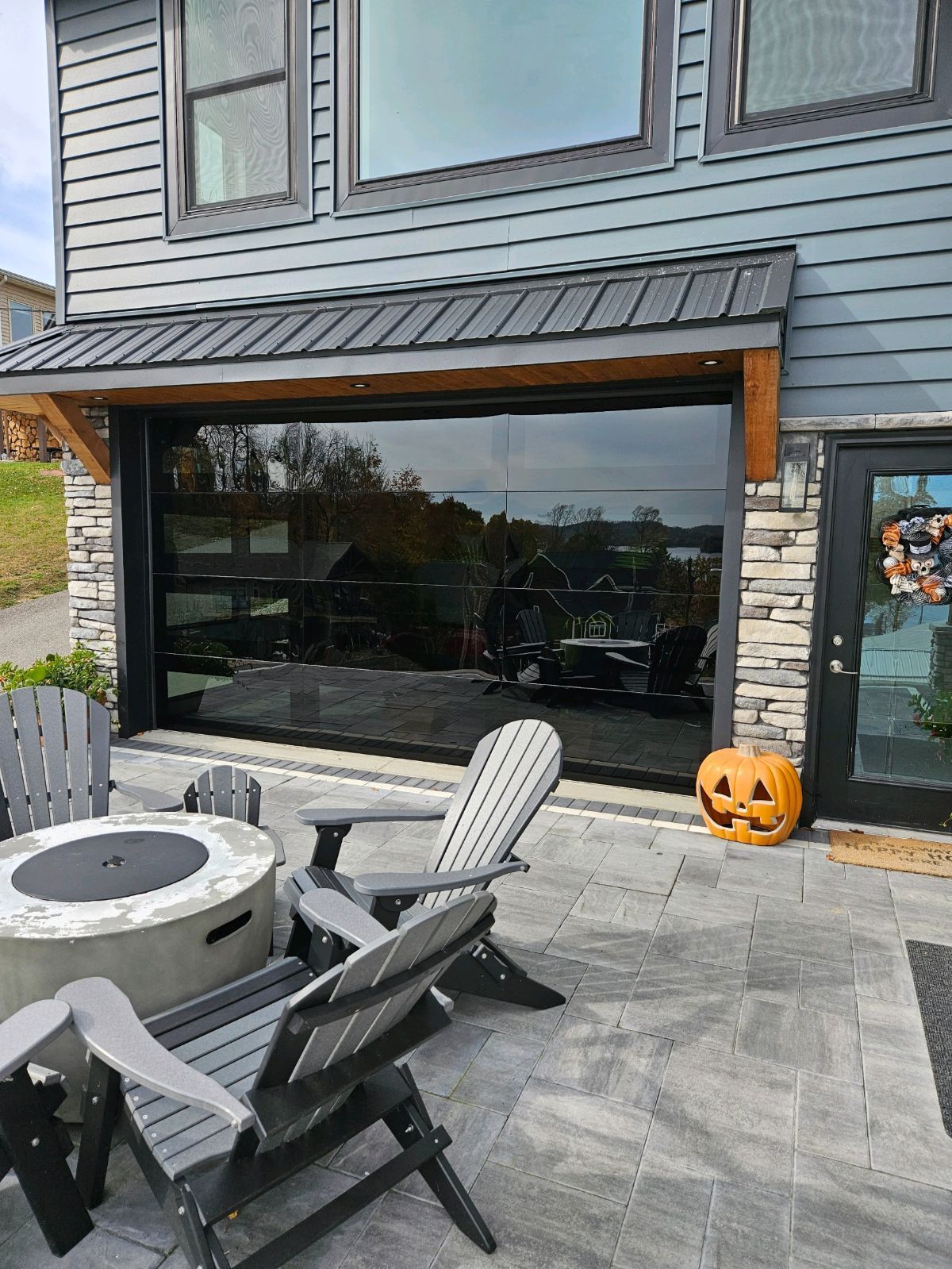 Modern home exterior with glass garage door, patio, Adirondack chairs, and a jack-o'-lantern.