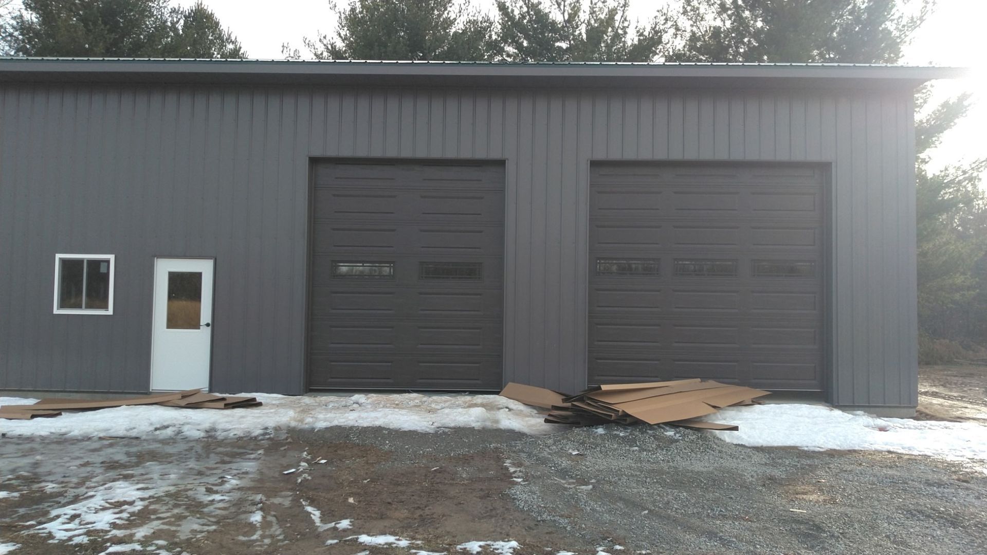 Gray metal building with two garage doors, a small door, and a window. Snow on the ground.