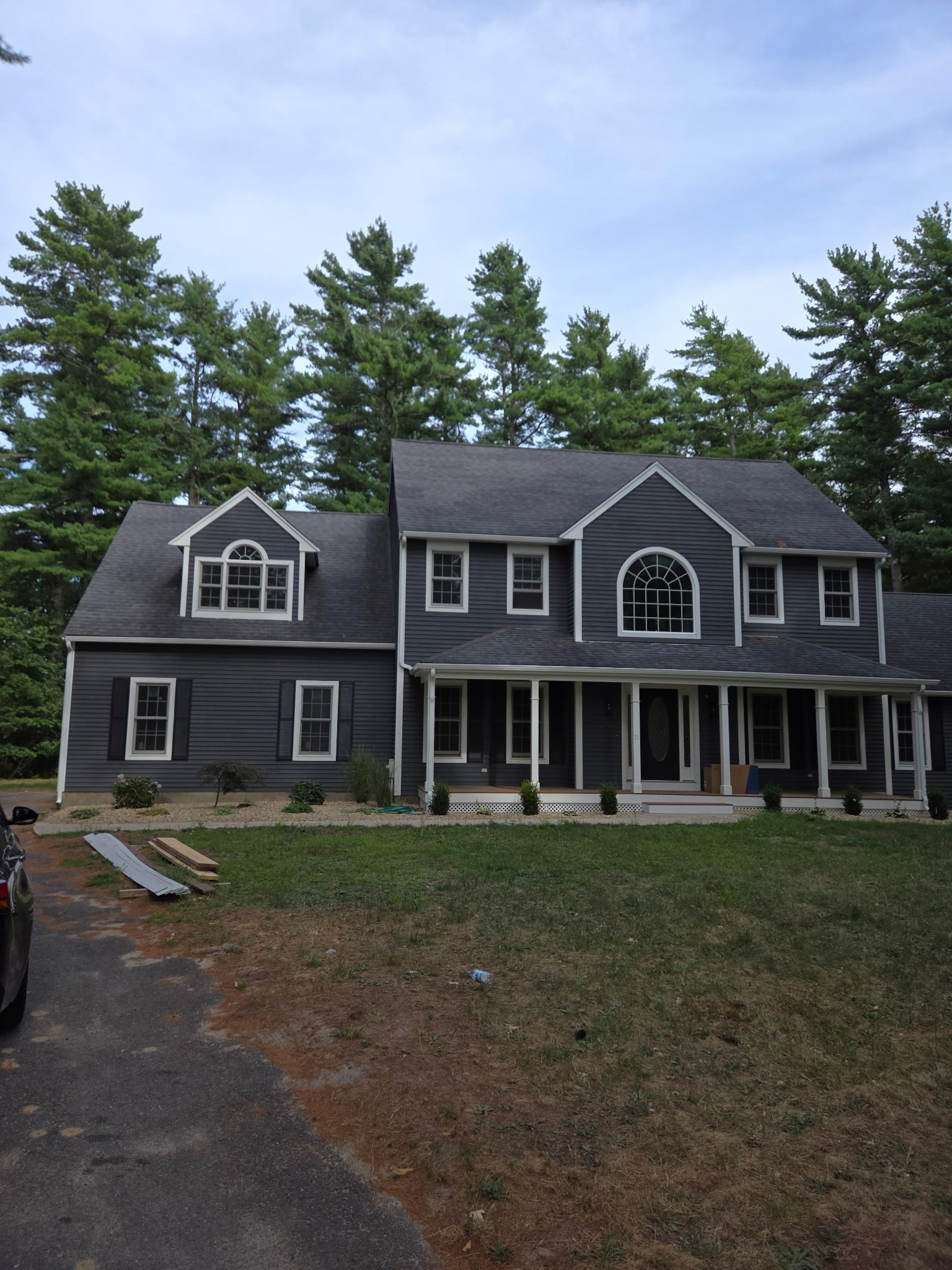 Gray two-story house with dark roof and trim, tall trees in the background, set on a lawn.