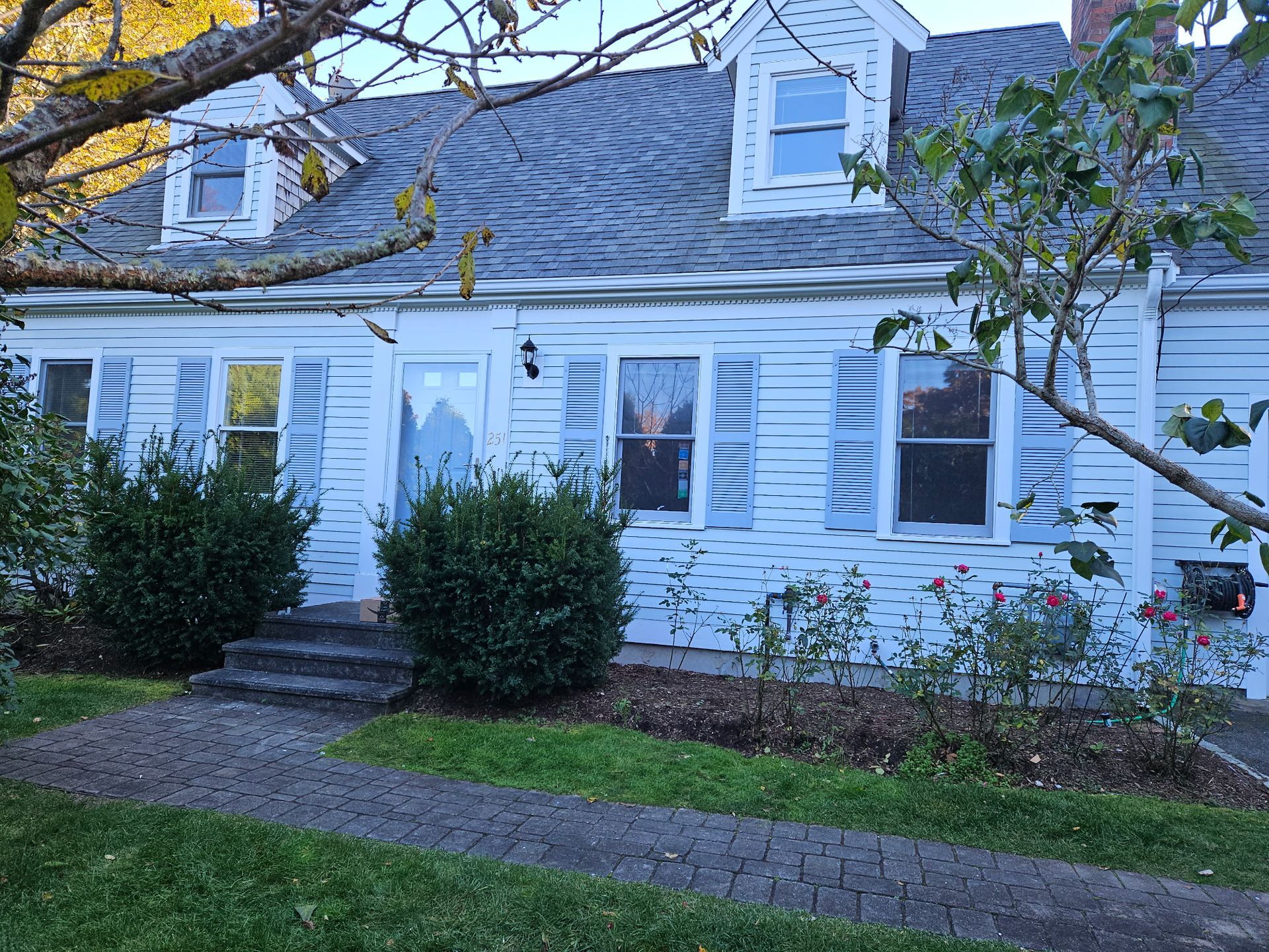 White house with blue shutters, front door, and three dormers. Green bushes and path lead to the entrance.