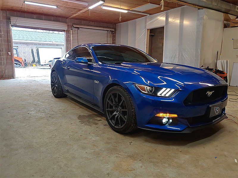 Blue Ford Mustang parked inside a garage with open doors, black wheels, and tinted windows.