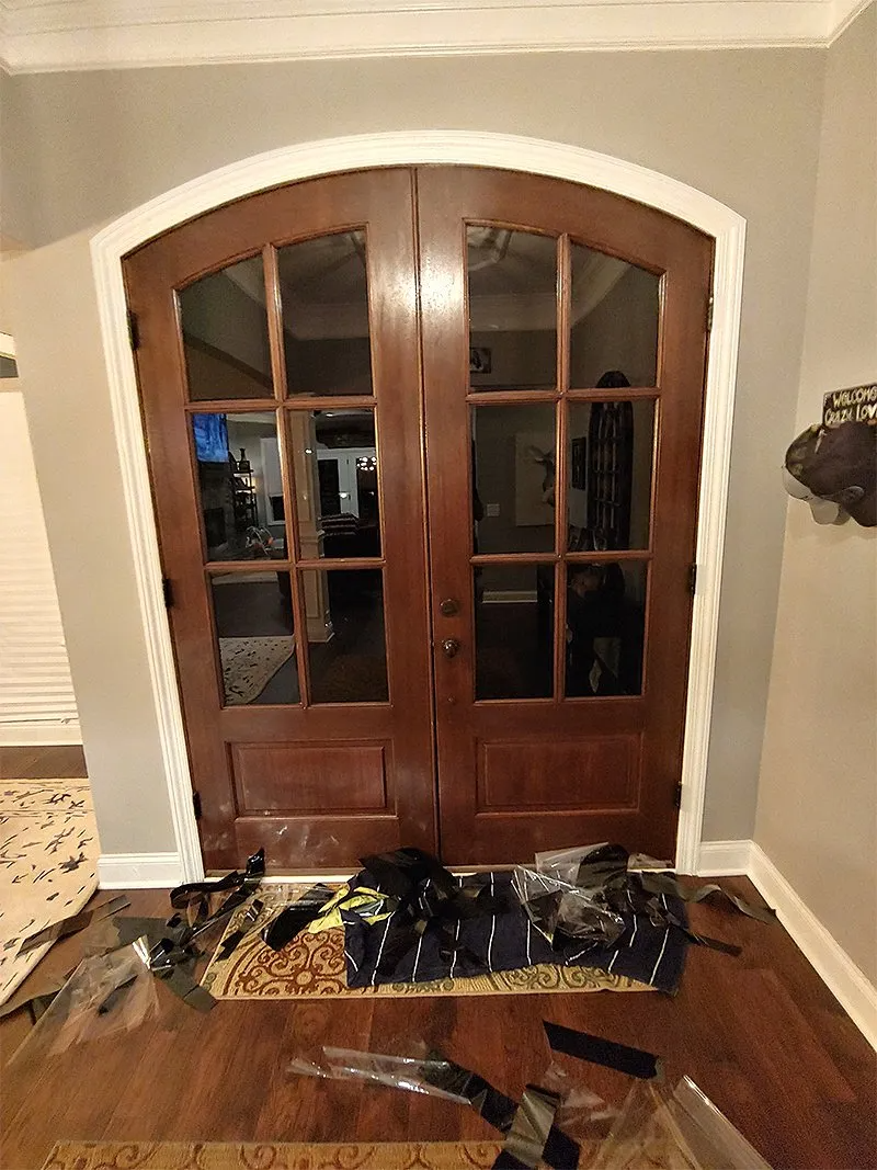 Brown arched double doors with glass panes, surrounded by white trim and a dark wooden floor. Debris in front.