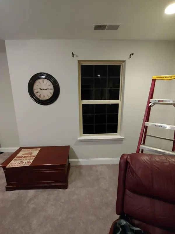 A room with a window, clock, table, recliner, and ladder. Light gray walls, brown furniture, and a neutral-colored carpet.