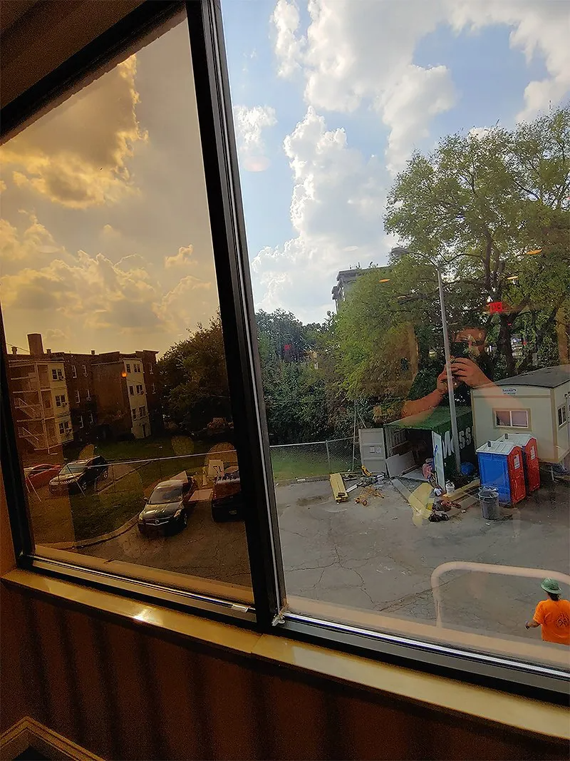 View through window: Cloudy sky, trees, building, cars, person holding pole near portable toilets.