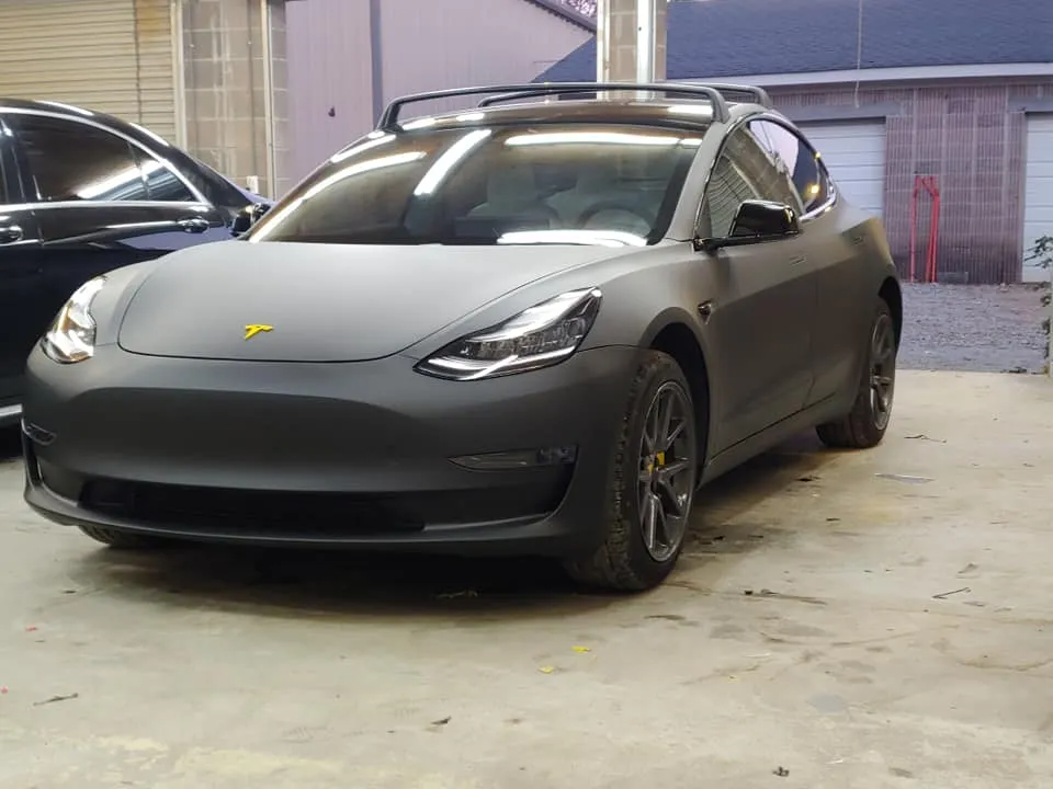 A matte black Tesla Model 3 with a roof rack parked inside a garage.