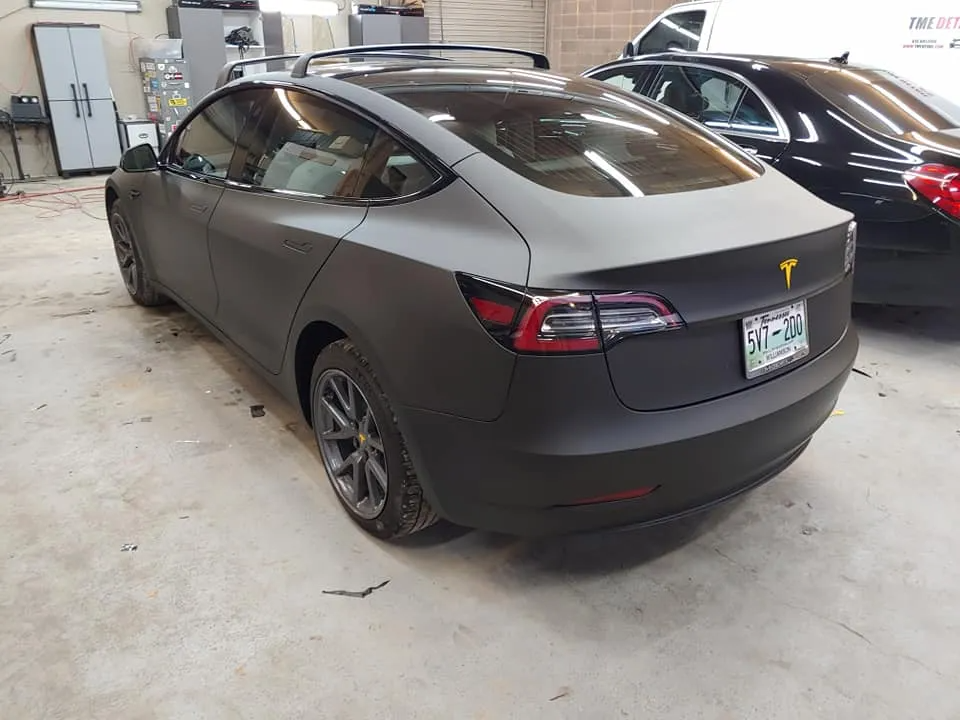 Dark gray Tesla car in a shop, with tinted windows and a yellow logo, angled from the back.