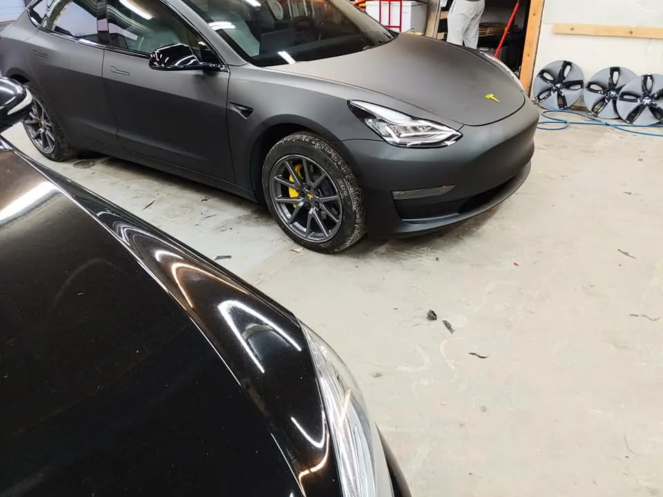 Dark gray Tesla and a black car next to it. The Tesla has yellow brake calipers, and is inside a garage.