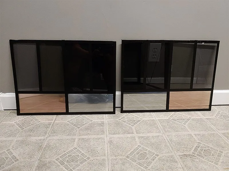 Two black framed decorative panels with glass, wood, and screen inserts, on a tiled floor against a gray wall.