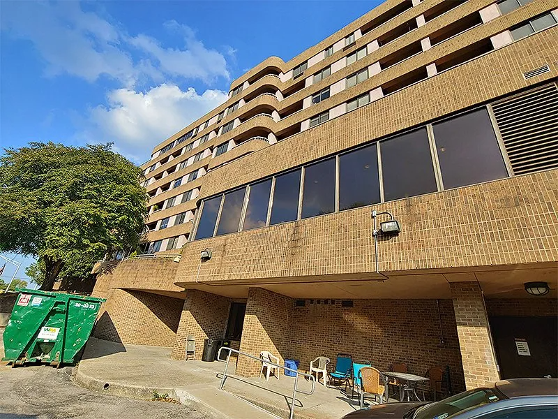 Brick high-rise building with several dark windows and a green dumpster in the foreground. Blue sky with clouds visible.