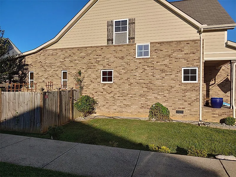Brick house with tan siding and small windows; a wooden fence and green grass are in the foreground.