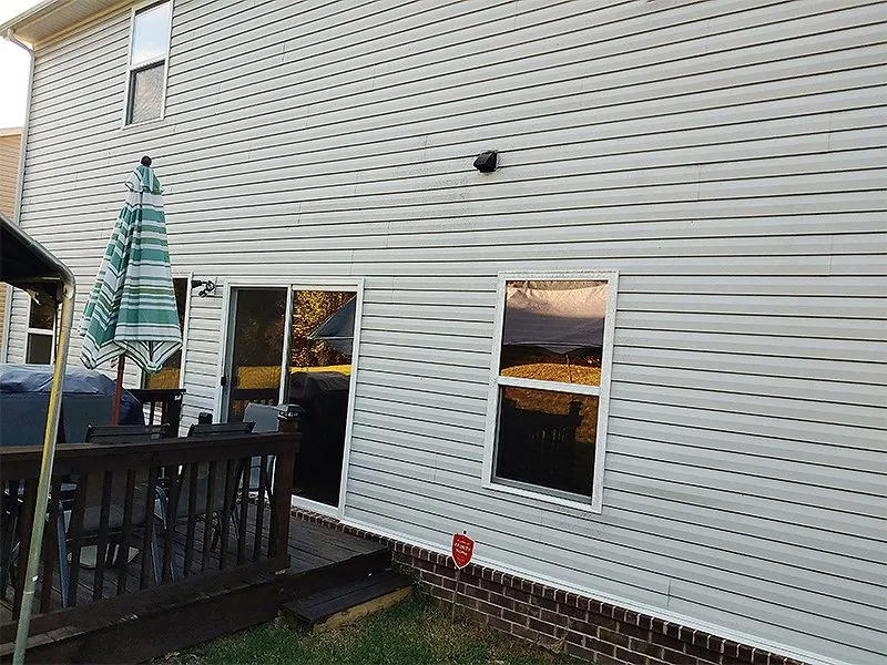 Back of a light gray vinyl-sided house with deck, sliding glass door, and window; setting sun reflecting.