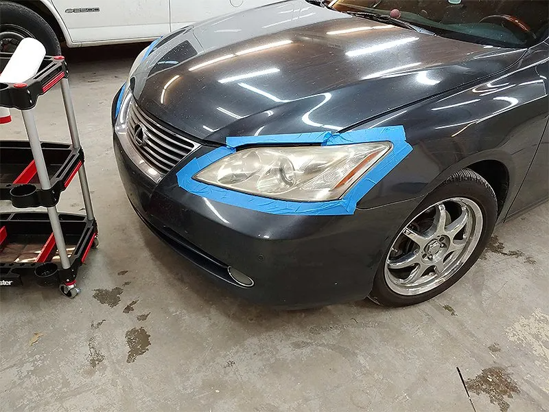 A gray car's headlights taped off, next to a detailing cart, in a shop.