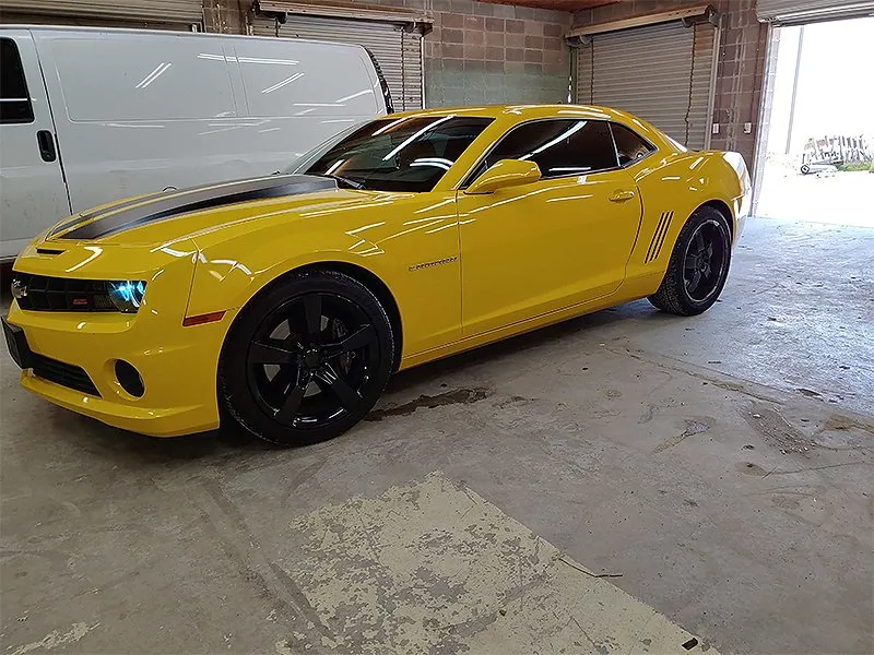 Yellow Camaro with black racing stripes and wheels parked in a garage.