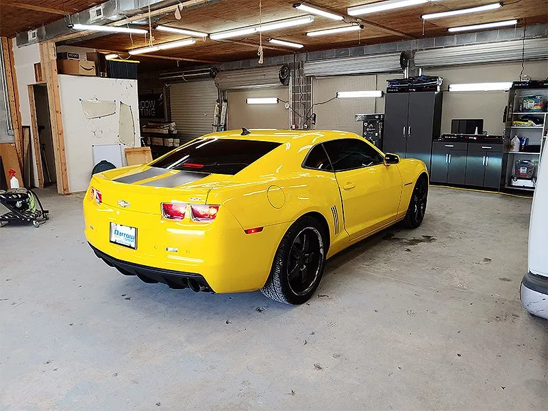 Yellow Camaro sports car in a garage, black racing stripes, black wheels.