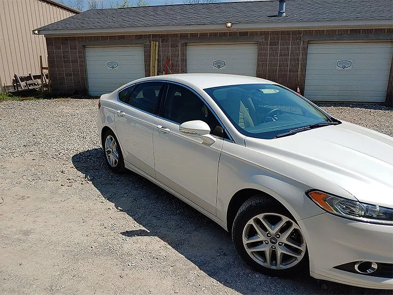 White Ford Fusion parked in front of a brick garage with three doors on a gravel driveway.