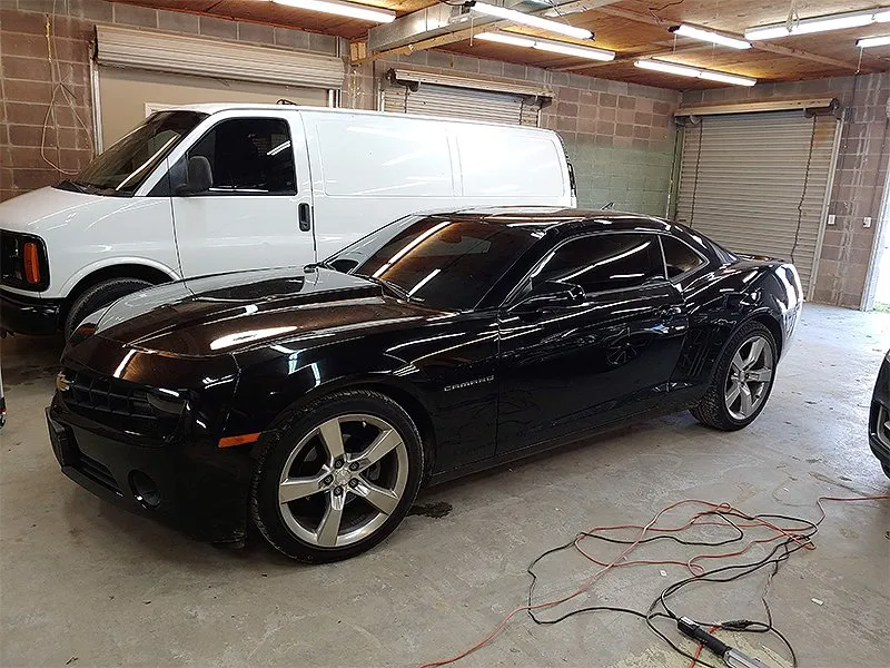Black car parked in a garage with a white van in the background, electrical cords on the floor.