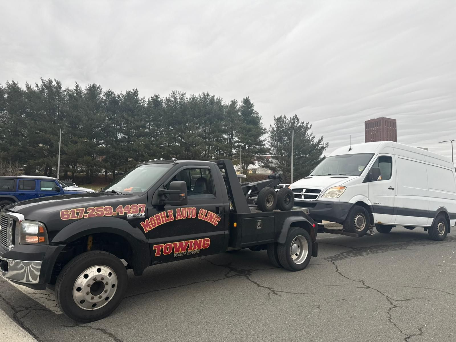 Black tow truck towing a white van, parked outdoors on a cloudy day.
