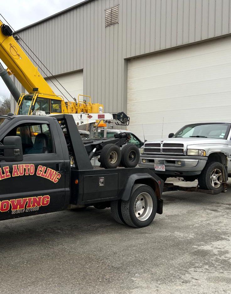 A silver pickup truck being towed by a black tow truck in front of a building. A yellow crane is visible in the background.