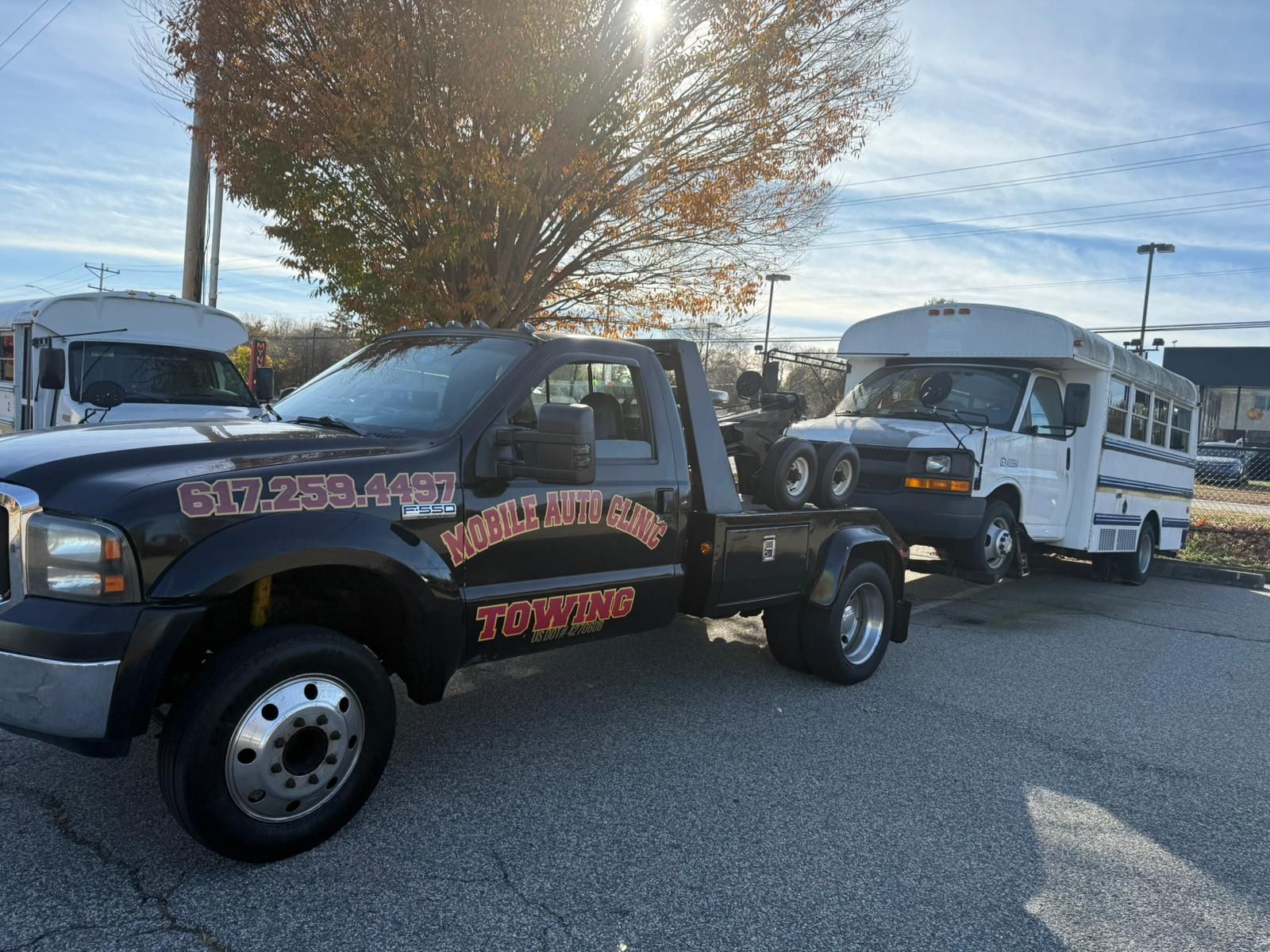 Black tow truck towing a white passenger bus on a paved lot with other vehicles visible.