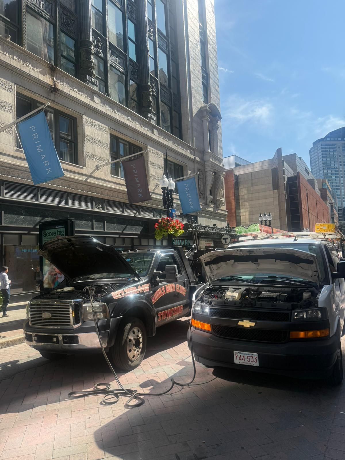 Two trucks with open hoods on a city street. One is black, the other gray, connected by cables.