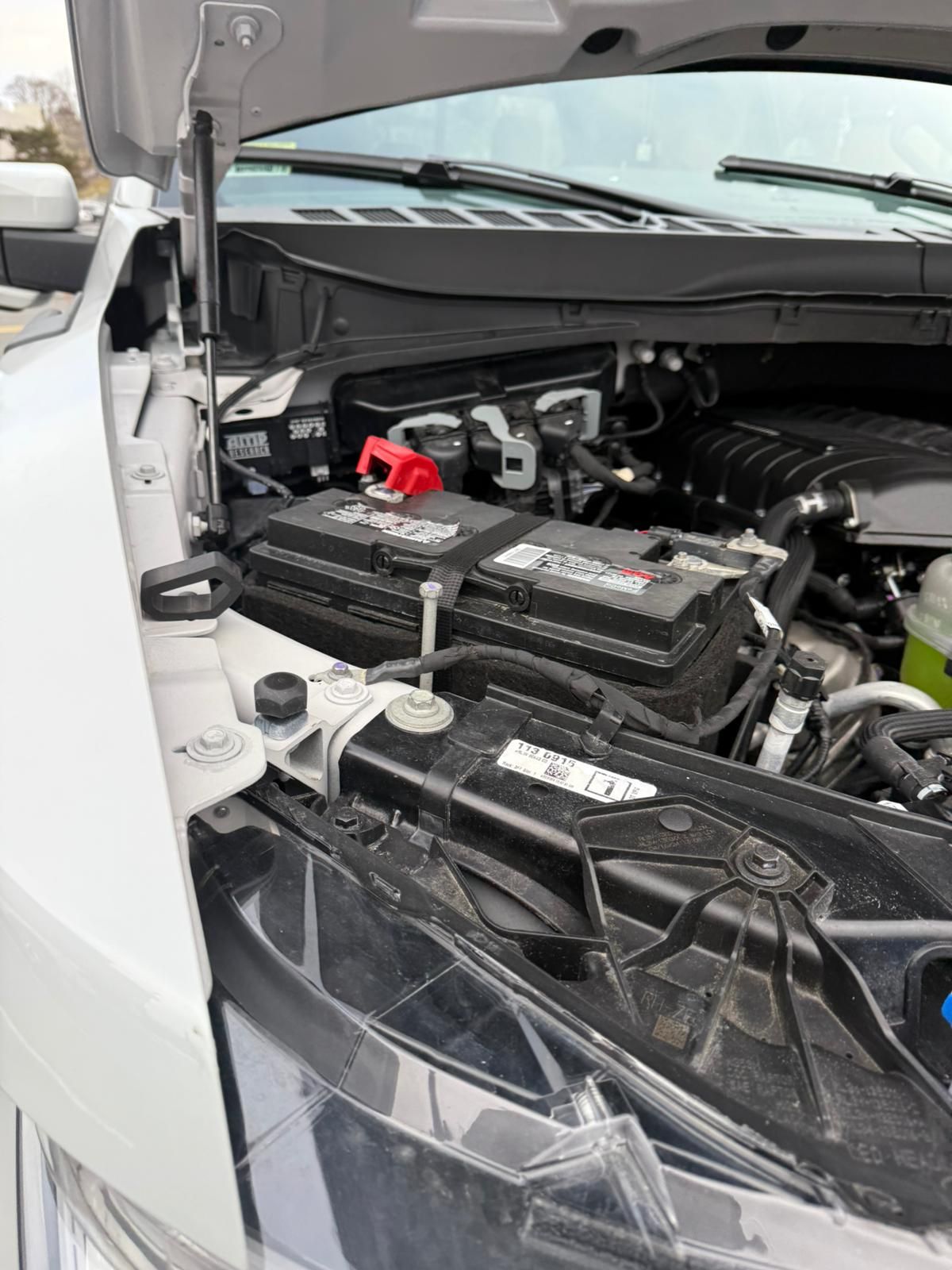 Open car hood revealing black battery with red positive terminal.