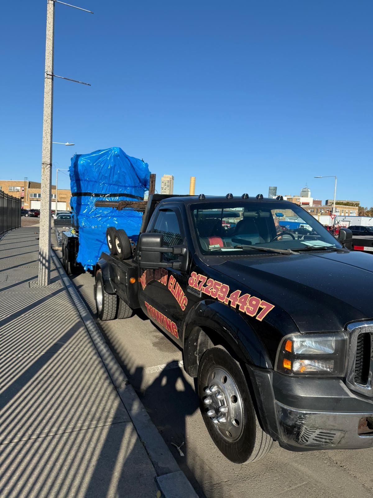 Black tow truck with a blue-covered load parked next to a sidewalk, under a blue sky.