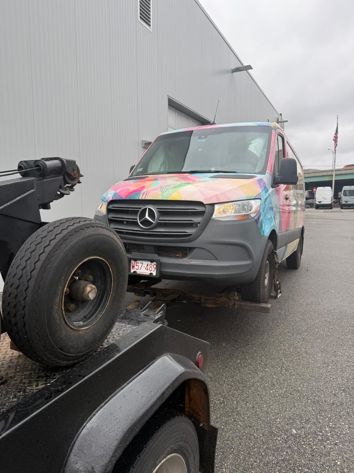 Mercedes van with colorful wrap being towed by a tow truck on a paved lot.