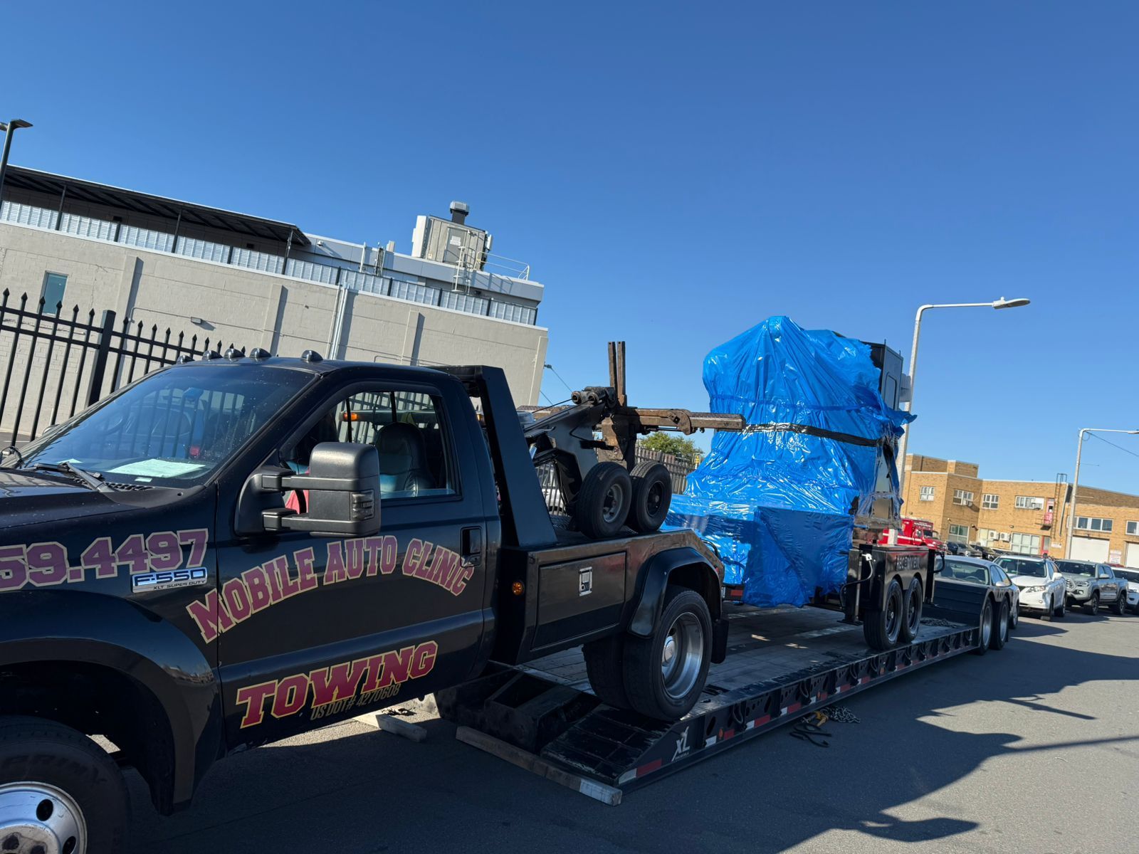 A black tow truck with a large, blue-wrapped object on its flatbed trailer on a city street.