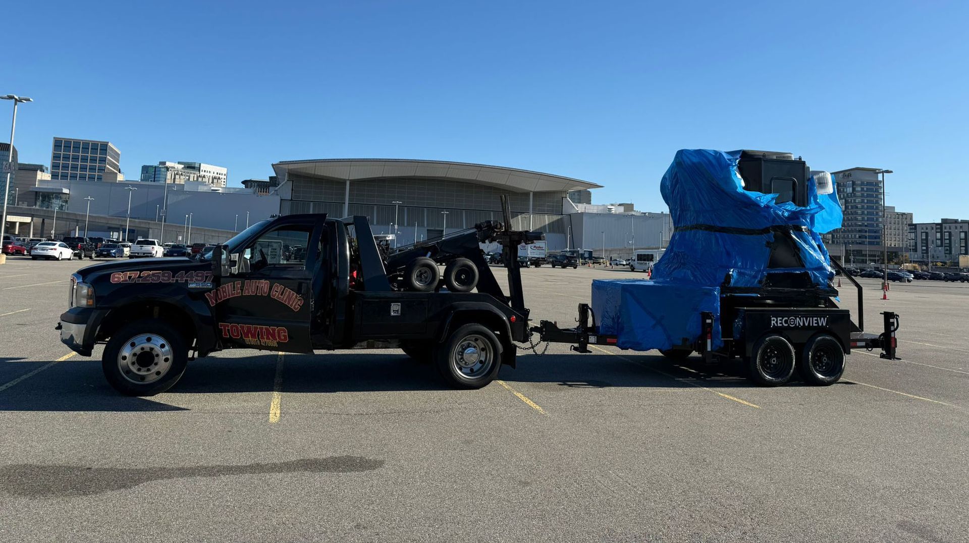 Black tow truck towing a trailer with large, blue-covered equipment in a parking lot on a sunny day.