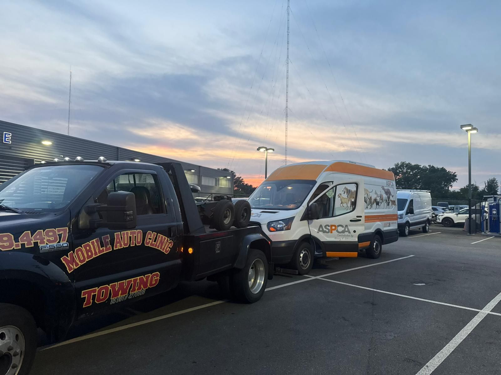 Tow truck towing an ASPCA van in a parking lot at dusk.