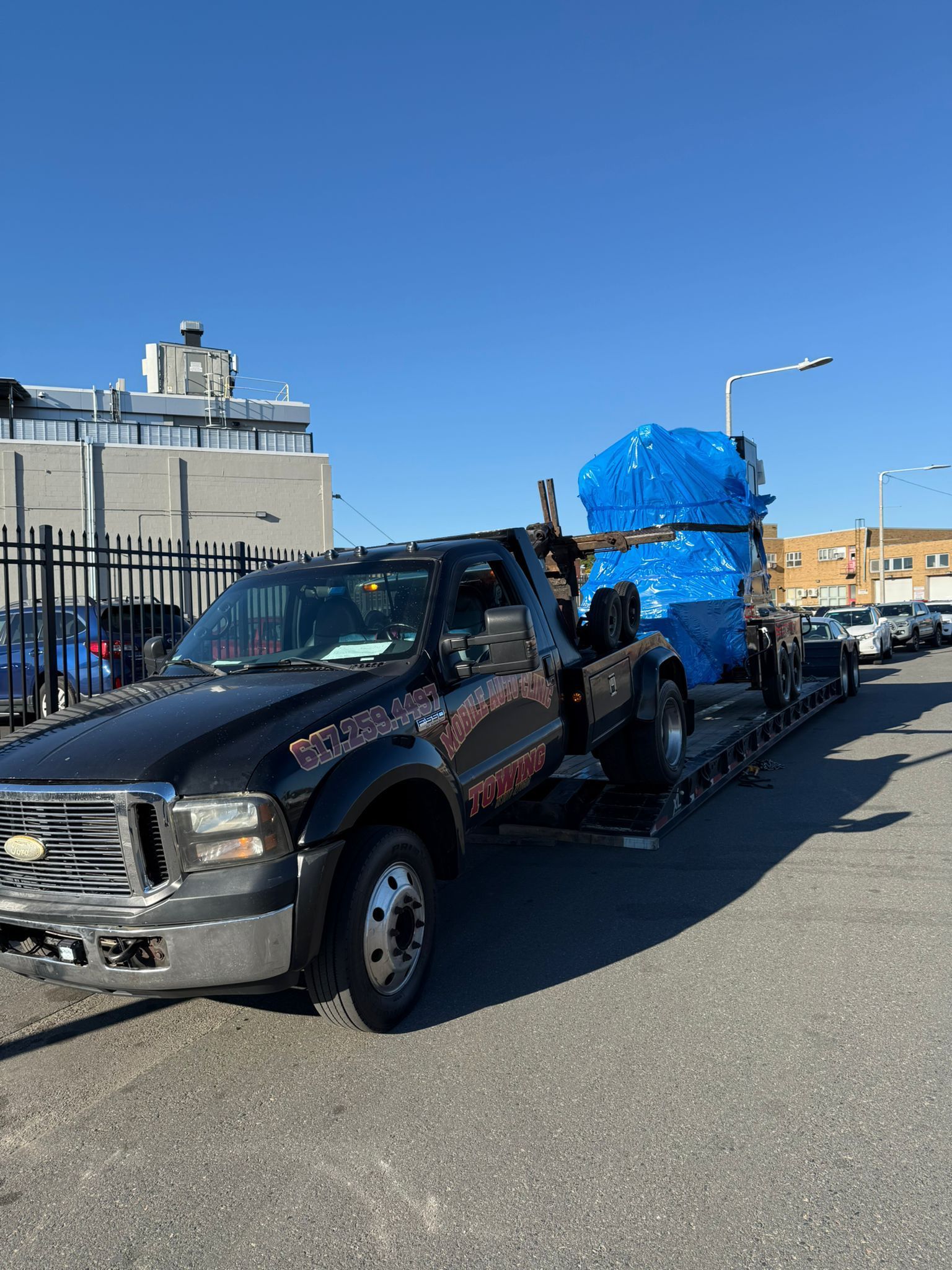 Black tow truck hauling a large blue-wrapped object on a flatbed trailer, parked on a city street.
