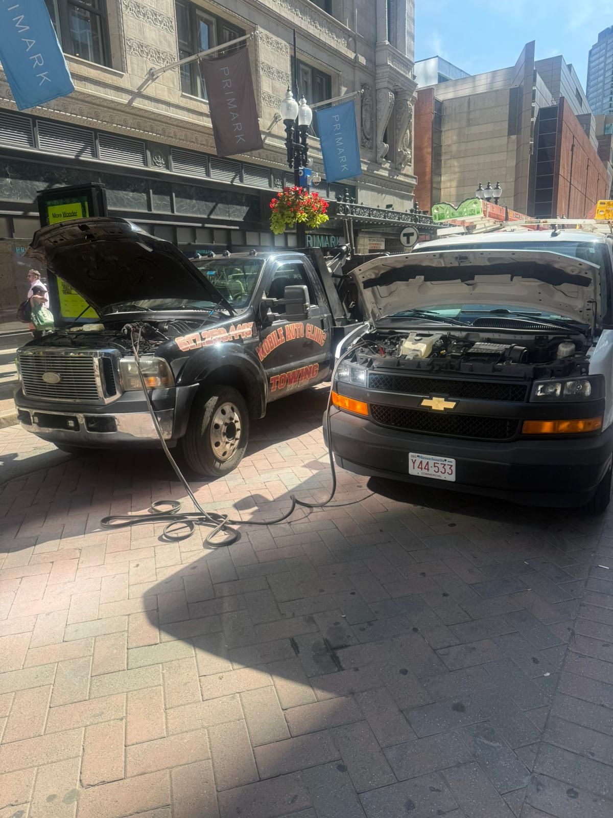 Two vehicles, a black truck and a white van, with open hoods, appear to be jump starting each other on a city street.