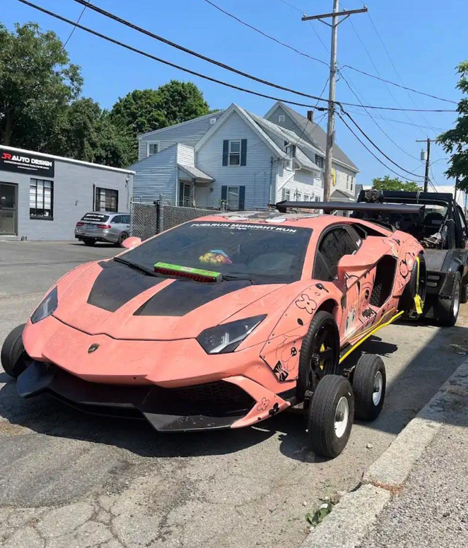 Pink, modified Lamborghini on oversized wheels, parked on a street next to a tow truck.