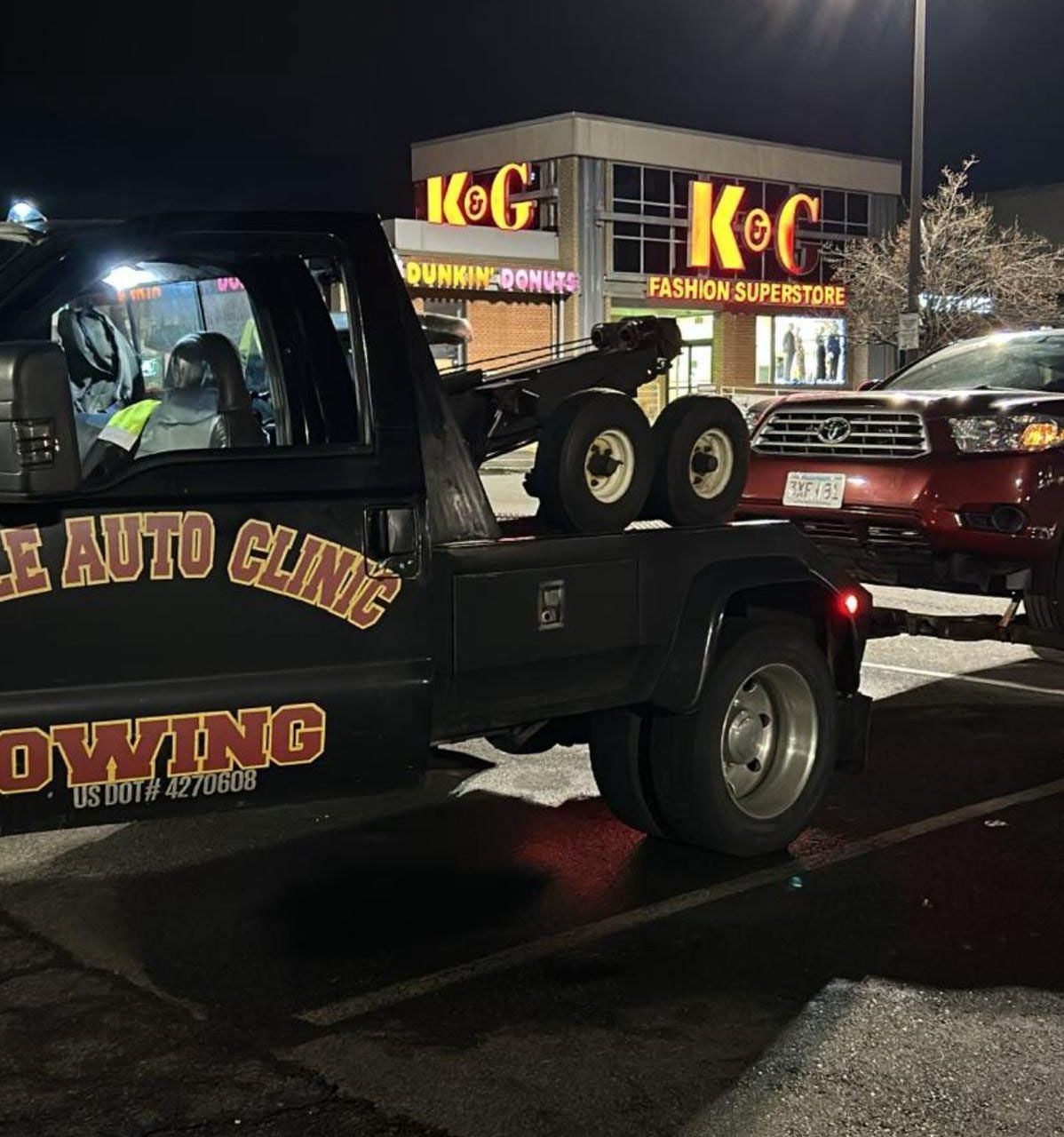 Tow truck towing a red car in a parking lot at night near a store with a lit up sign.