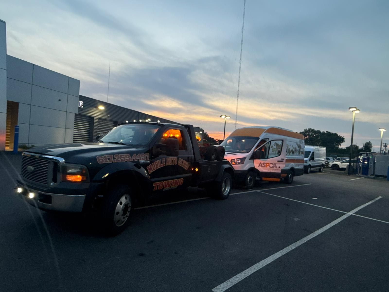 Tow truck towing a van in a parking lot at dusk. Black truck, white and orange van, cloudy sky.