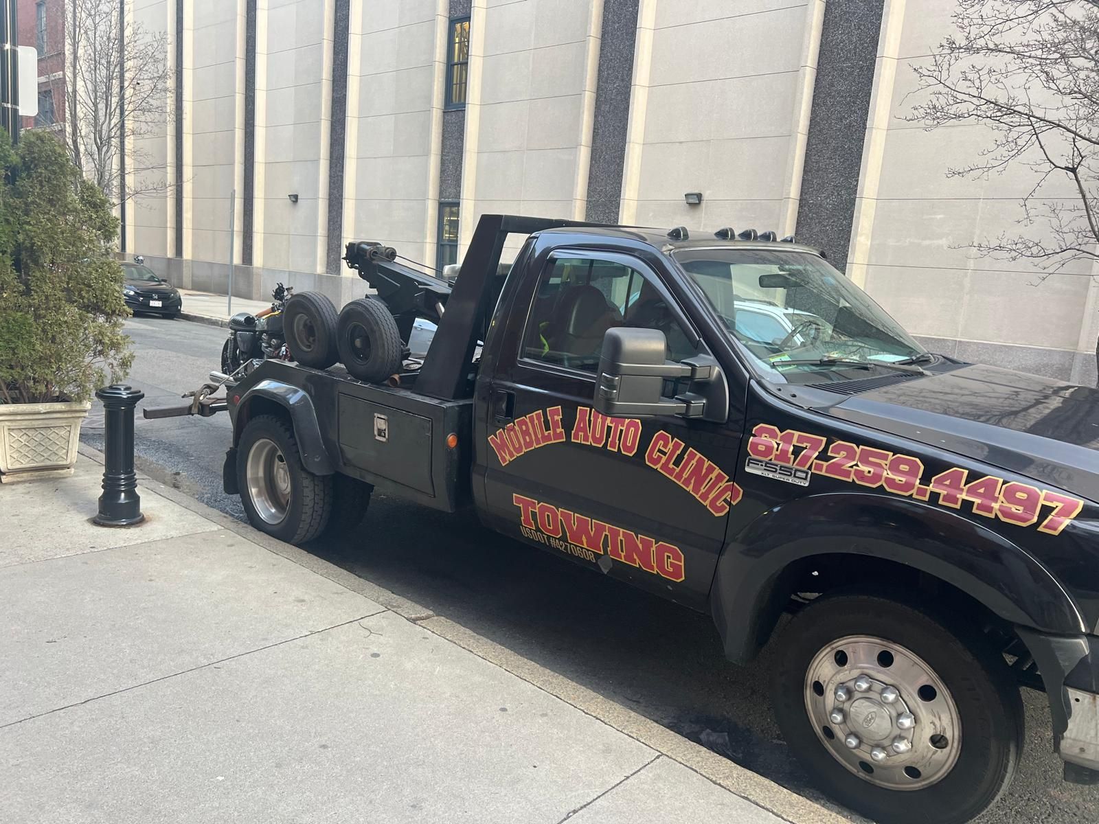 Black tow truck parked on a sidewalk, towing another vehicle. Building in the background.