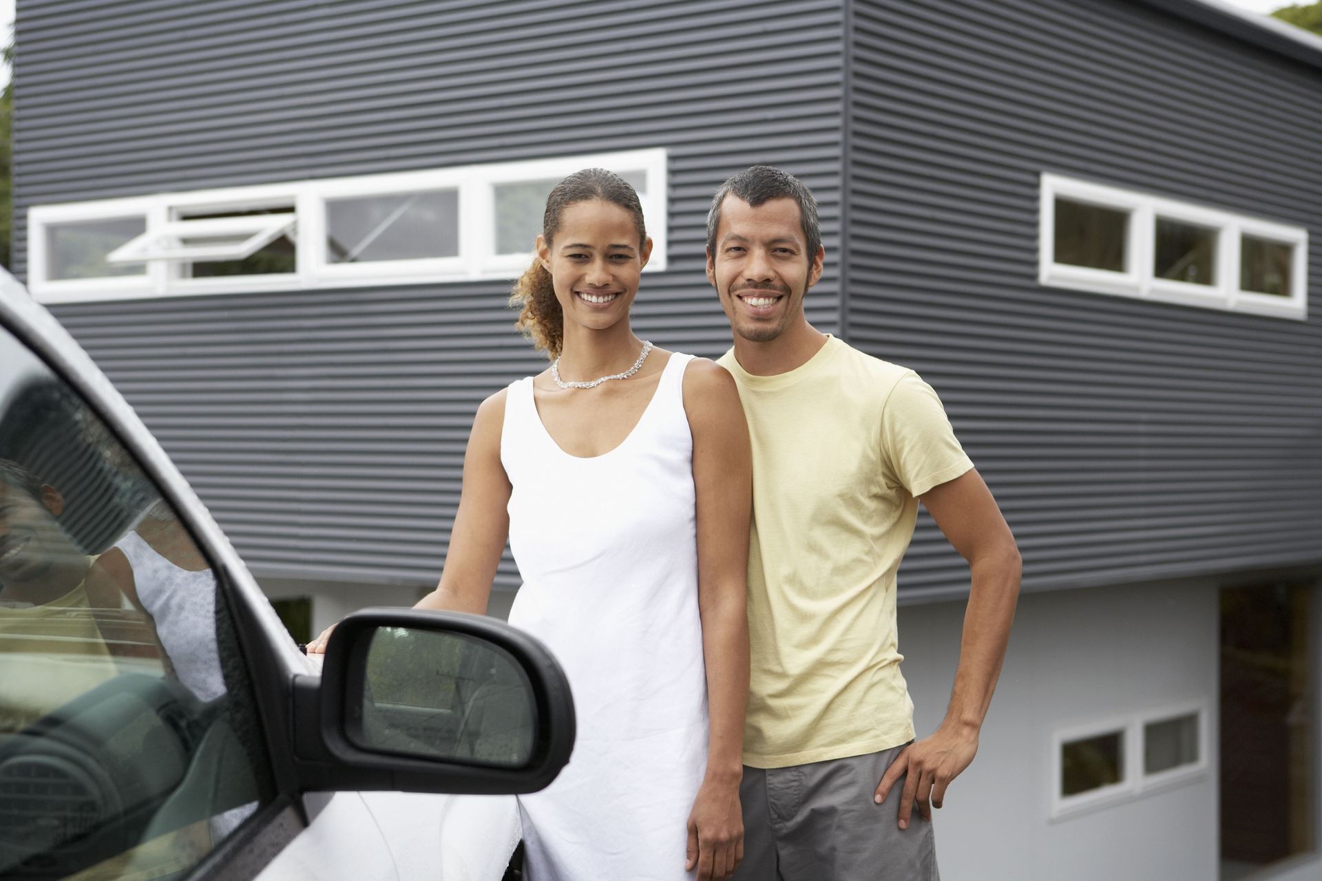 Couple smiling in front of a modern grey house next to a white car.