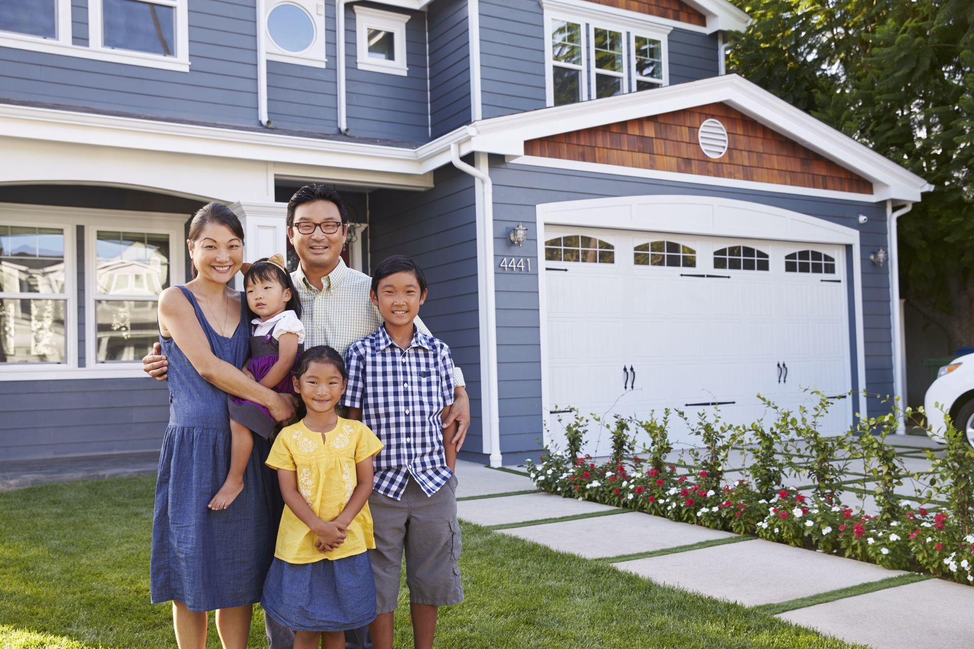 Family of five smiling in front of their blue house with a white garage. Green lawn, sunny day.