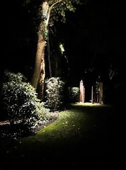 Nighttime garden path illuminated by spotlights, bordered by hedges and trees.