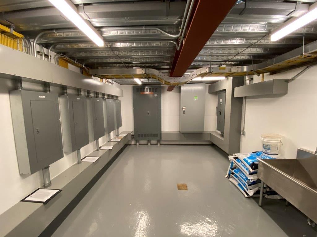 Interior of a utility room with several gray electrical panels along the left wall, a central walkway, and various equipment.