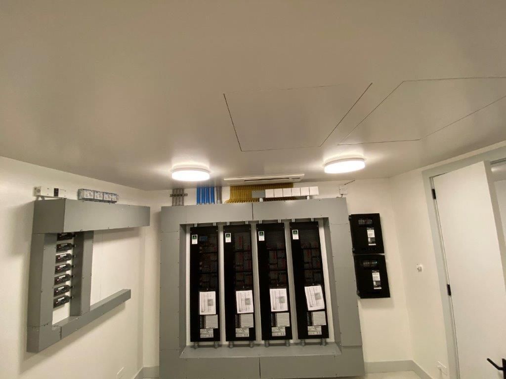 Electrical control panel room with gray panels, white walls, and two ceiling lights.