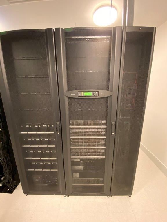 Three dark server racks in a brightly lit room. The middle rack has a digital display.